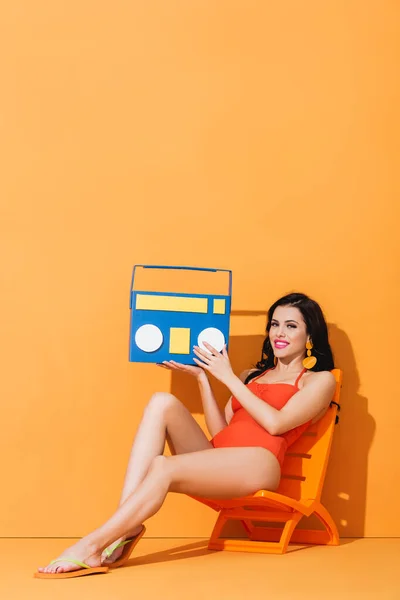 Happy woman in swimsuit holding paper boombox while sitting on deck chair on orange — Stock Photo