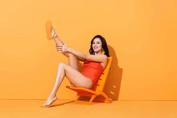 Cheerful woman in swimsuit touching leg and sitting on deck chair on orange — Stock Photo