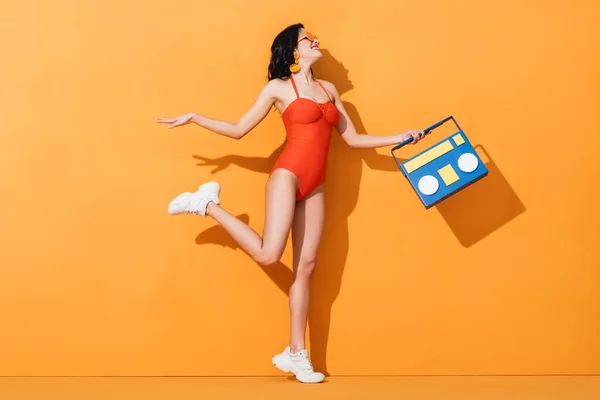 Happy woman in sneakers, sunglasses and bathing suit holding paper cut boombox on orange — Stock Photo