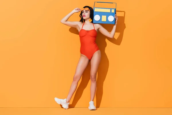 Femme élégante en baskets et maillot de bain touchant lunettes de soleil tout en tenant le papier coupe boombox sur orange — Photo de stock