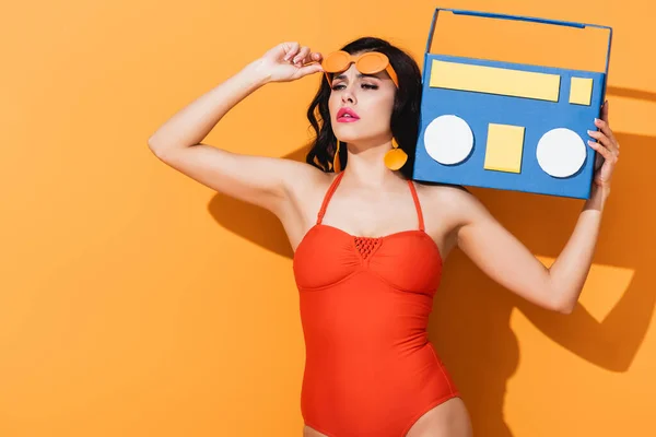 Stylish woman in bathing suit touching sunglasses while holding paper cut boombox on orange — Stock Photo