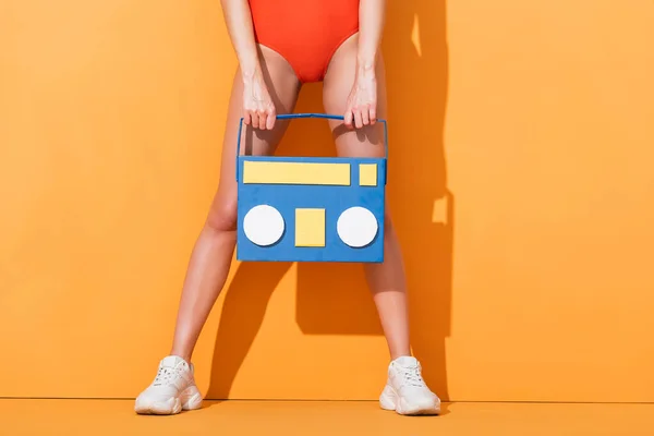 Vue recadrée de la jeune femme en baskets et maillot de bain tenant le papier coupé boombox sur orange — Photo de stock