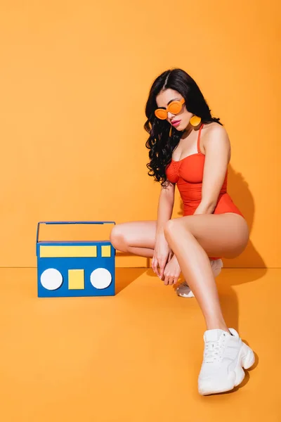 Stylish woman in sneakers, bathing suit and sunglasses sitting near paper cut boombox on orange — Stock Photo