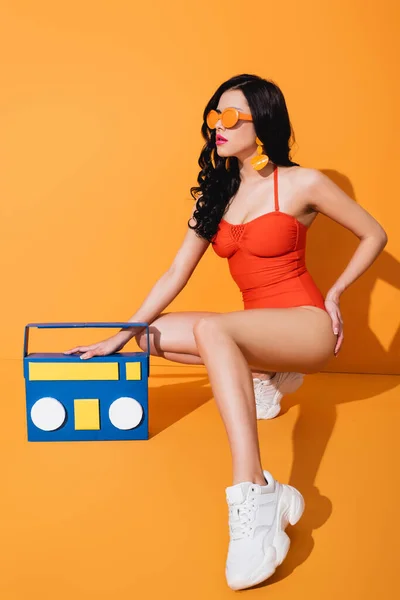 Chica con estilo en zapatillas de deporte, traje de baño y gafas de sol sentado cerca de la caja de boom artesanal de papel en naranja - foto de stock