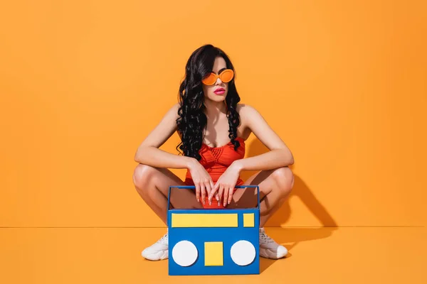 Jeune femme à la mode en baskets, maillot de bain et lunettes de soleil assis près de la boombox de papier coupé sur orange — Photo de stock