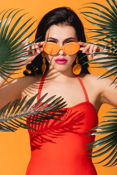 Young and stylish woman touching sunglasses near green palm leaves isolated on orange — Stock Photo