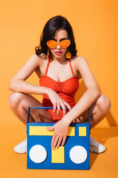 Mujer joven con estilo en zapatillas de deporte, traje de baño y gafas de sol sentados cerca de la caja de resonancia de papel en naranja - foto de stock