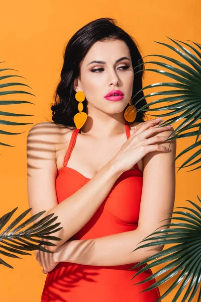 Young woman touching shoulder near green palm leaves isolated on orange — Stock Photo