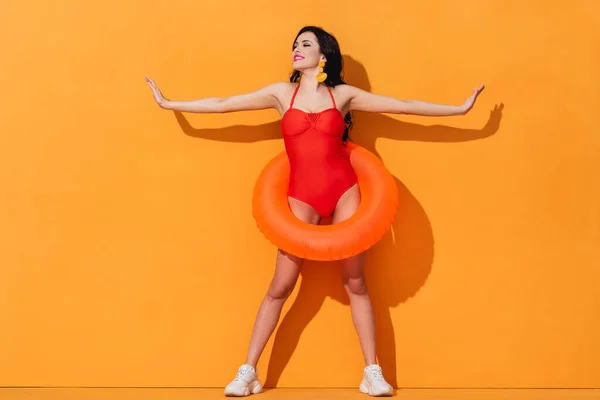 Счастливая девушка в купальнике с надувным кольцом и протянутыми руками, стоящая на оранжевом — стоковое фото