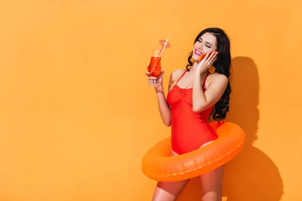 Fille gaie en maillot de bain avec anneau gonflable tenant cocktail et parler sur smartphone sur orange — Photo de stock