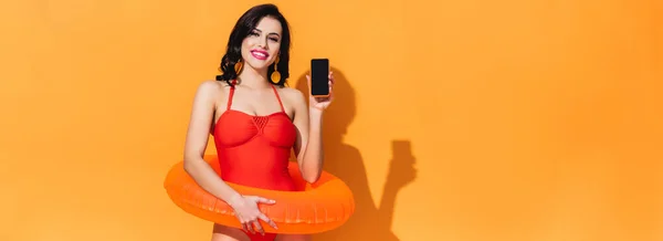 Panoramakonzept einer glücklichen Frau im Badeanzug mit aufblasbarem Ring, der Smartphone mit leerem Bildschirm auf orange hält — Stockfoto