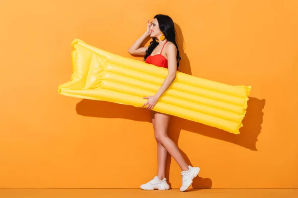 Side view of cheerful girl in swimsuit and sneakers holding inflatable mattress on orange — Stock Photo