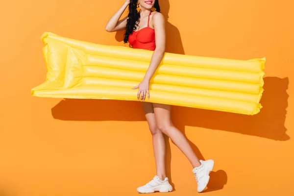 Vista recortada de chica feliz en traje de baño y zapatillas de deporte con colchón inflable en naranja - foto de stock