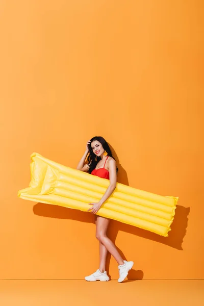 Happy woman in swimsuit and sneakers holding inflatable mattress and touching hair on orange — Stock Photo