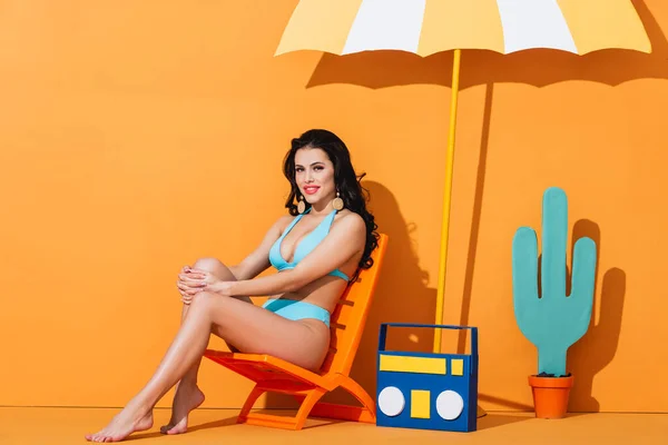 Heureuse jeune femme en maillot de bain assis sur la chaise longue près de la boombox en papier, cactus et parapluie sur orange — Photo de stock