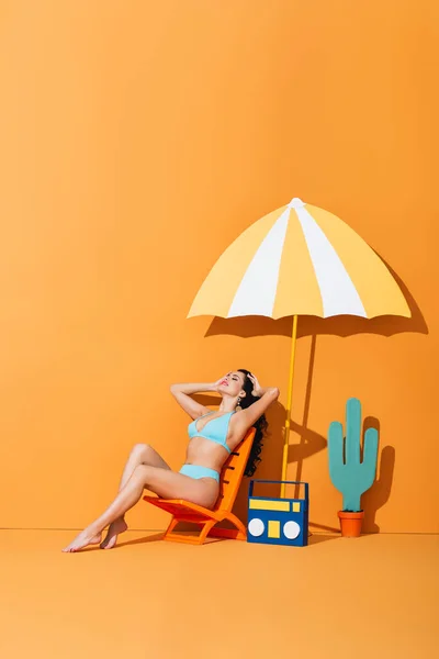 Femme heureuse avec les yeux fermés assis sur la chaise longue près de boombox en papier, cactus et parapluie sur orange — Photo de stock