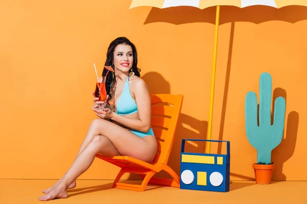 Femme heureuse en maillot de bain assis sur la chaise longue près de la boombox en papier, cactus et parapluie tout en tenant un cocktail sur orange — Photo de stock