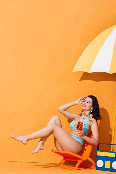 Cheerful woman in swimwear sitting on deck chair near paper umbrella and boombox while holding cocktail on orange — Stock Photo