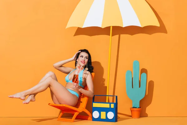 Mujer sonriente en traje de baño sentado en la silla de cubierta cerca de la caja de resonancia de papel, cactus y paraguas mientras que la celebración de cóctel en naranja - foto de stock