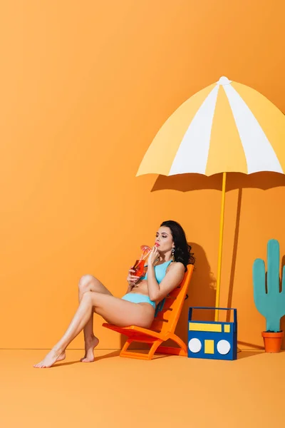 Femme en maillot de bain assis sur la chaise longue près de la boombox en papier, cactus et parapluie tout en buvant un cocktail sur orange — Photo de stock