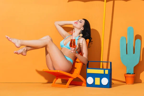 Attraente donna in costume da bagno seduto sulla sedia a sdraio vicino alla scatola di carta e cactus mentre tiene cocktail su arancione — Foto stock