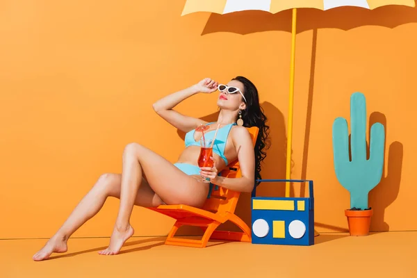 Fille élégante en lunettes de soleil et maillot de bain assis sur la chaise longue près de la boombox en papier, cactus et parapluie tout en tenant un cocktail sur orange — Photo de stock