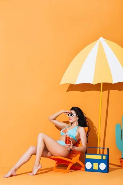 Стильная женщина в солнечных очках и купальнике сидит на шезлонге возле бумажного бумбокса и зонтика, держа коктейль на апельсине — стоковое фото