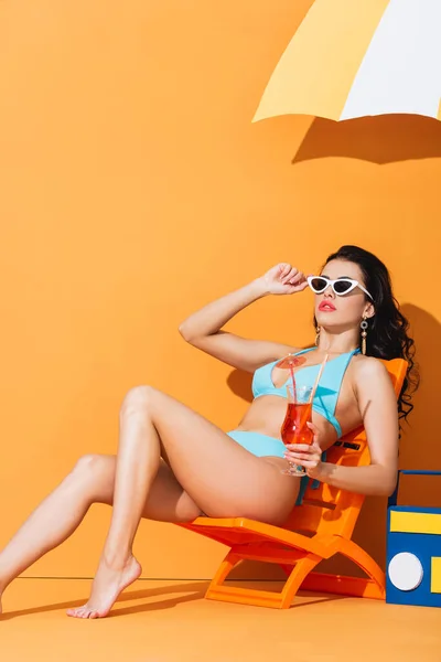 Trendfrau in Sonnenbrille und Badeanzug sitzt auf Liegestuhl neben Papierbox und Regenschirm und hält Cocktail auf Orange — Stockfoto