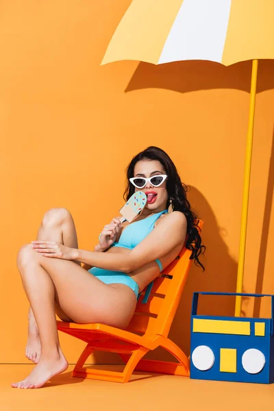 Mujer de moda en gafas de sol y trajes de baño sentado en la silla de cubierta cerca de la caja de boom de papel y paraguas mientras lame helado en naranja - foto de stock