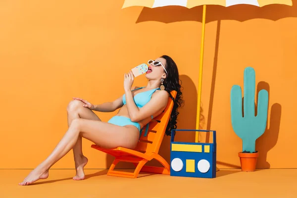 Mujer de moda en gafas de sol y trajes de baño sentado en la silla de cubierta cerca de la caja de boom y paraguas mientras lame helado de papel en naranja - foto de stock