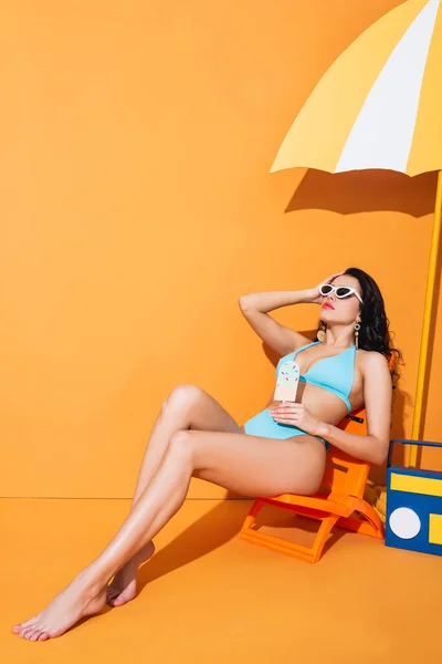 Femme à la mode en lunettes de soleil et maillots de bain assis sur la chaise longue près de la boombox en papier et parapluie tout en tenant la crème glacée sur orange — Photo de stock