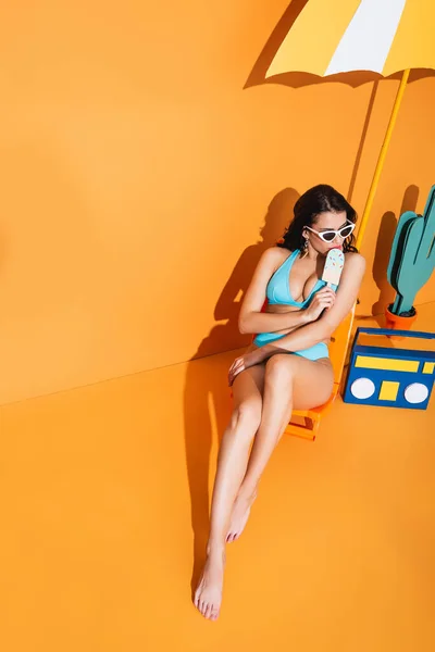 Vue grand angle de la femme à la mode dans les lunettes de soleil et maillots de bain assis sur la chaise longue près de la boombox en papier et le parapluie tout en tenant la crème glacée sur orange — Photo de stock
