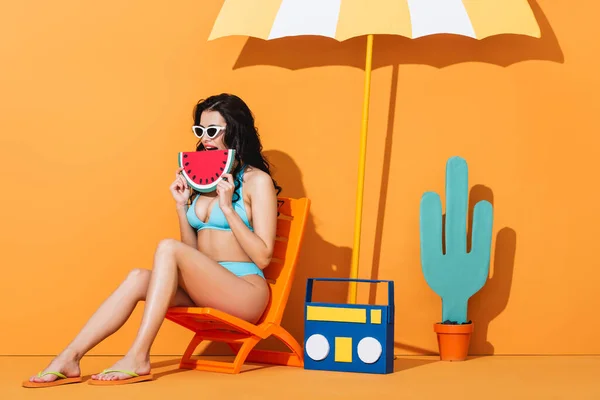 Молодая женщина в солнечных очках и купальнике сидит на шезлонге возле бумбокса и зонтика, держа в руках бумажный арбуз на апельсине — стоковое фото