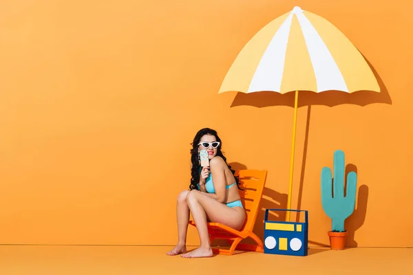 Happy woman in sunglasses and swimsuit sitting on deck chair near boombox and umbrella while holding paper ice cream on orange — Stock Photo