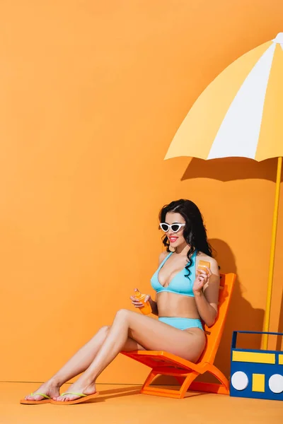 Happy woman in sunglasses and swimsuit sitting on deck chair near paper boombox, cactus and umbrella while applying sunscreen on orange — Stock Photo