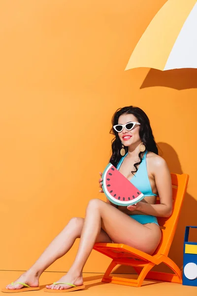 Cheerful woman in sunglasses and swimsuit sitting on deck chair near boombox and umbrella while holding paper watermelon on orange — Stock Photo