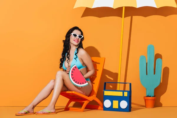 Cheerful girl in sunglasses and swimsuit sitting on deck chair near boombox, cactus and umbrella while holding paper watermelon on orange — Stock Photo