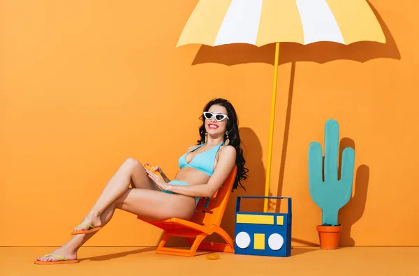 Счастливая девушка в солнечных очках и купальнике, сидящая на шезлонге возле бумажного бумбокса, кактуса и зонтика при нанесении солнцезащитного крема на апельсин — стоковое фото