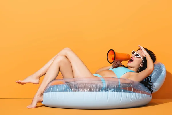 Young woman in swimsuit lying on inflatable ring and screaming in megaphone on orange — Stock Photo