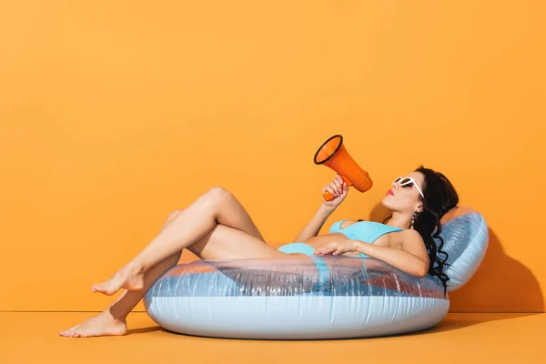 Barefoot woman in sunglasses and swimwear lying on inflatable ring and holding megaphone on orange — Stock Photo