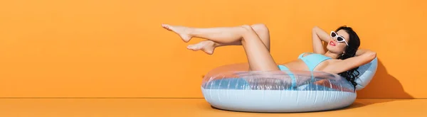 Panoramic crop of barefoot woman in sunglasses and swimwear lying on inflatable ring on orange — Stock Photo