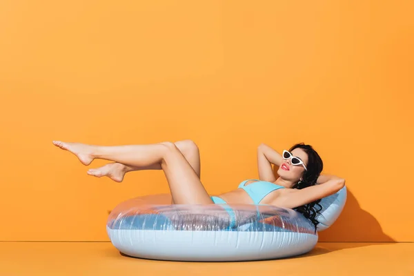 Весела жінка в сонцезахисних окулярах і купальнику лежить на надувному кільці на помаранчевому — стокове фото
