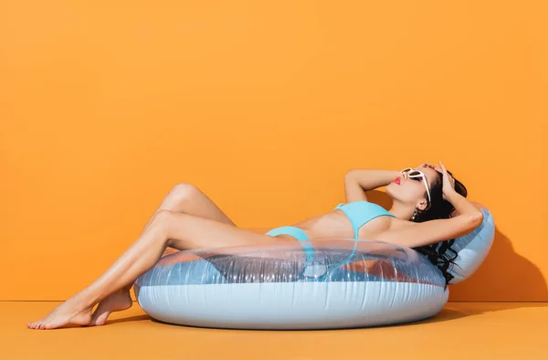 Stylish woman in sunglasses and swimwear lying on inflatable ring on orange — Stock Photo