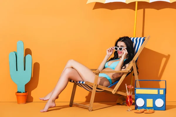 Happy woman in swimwear touching sunglasses and sitting on deck chair near cocktail, flip flops, paper boombox and cactus on orange — Stock Photo