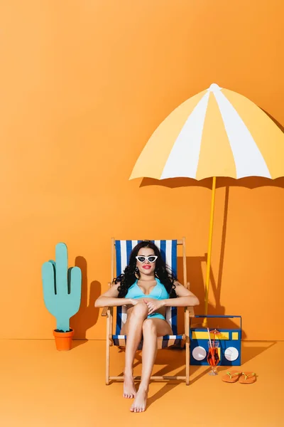Glückliche Frau in Badebekleidung und Sonnenbrille, die auf einem Liegestuhl in der Nähe von Cocktail, Flip Flops, Papierboombox und Kaktus auf Orange sitzt — Stockfoto