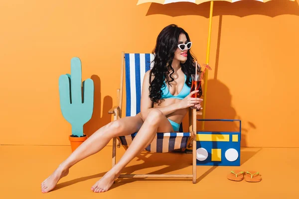 Femme heureuse en maillots de bain et lunettes de soleil assis sur la chaise longue et tenant cocktail près de tongs, boombox en papier et cactus sur orange — Photo de stock