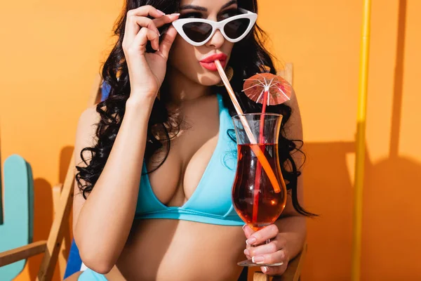 Young woman in swimwear touching sunglasses and drinking cocktail on orange — Stock Photo