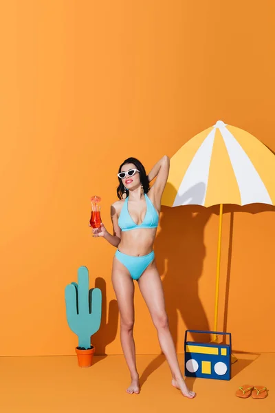 Happy woman in swimwear and sunglasses holding cocktail near paper umbrella, boombox and cactus on orange — Stock Photo