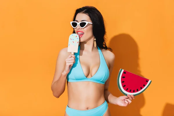 Mujer joven en traje de baño lamiendo helado de papel y sosteniendo sandía en naranja — Stock Photo