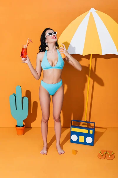 Donna felice in costume da bagno e occhiali da sole applicare la protezione solare e tenere cocktail vicino ombrello di carta, boombox e cactus su arancione — Foto stock